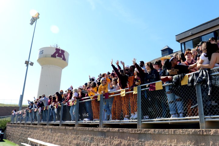 An older professor wearing a mask leans over a student's shoulder, gesturing toward a computer screen displaying lines of code. The professor appears to be providing guidance or explaining something, while the student listens attentively. The room has a soft, dim light.